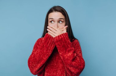 Bir kadın mavi arka planda duruyor. O, elleriyle ağzını kapatır ve bir şaşkınlık ifadesi gösterir. Uzun kahverengi saçları var ve kırmızı bir kazak giyiyor..