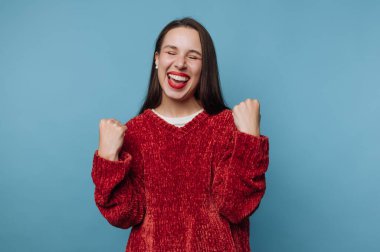 Bir kadın mavi bir duvarın önünde duruyor, geniş bir gülümsemeyle. Uzun saçları var ve kırmızı bir kazak giyiyor. Mutluluktan elleri kalkıyor, sıradan bir ortamda heyecanını gösteriyor..