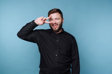 Mavi bir duvarın önünde duran bir adam, bir eliyle göz kırparken barış işareti yapıyor. Siyah bir gömlek giyiyor ve sıradan bir anda neşeli bir ifade sergiliyor..