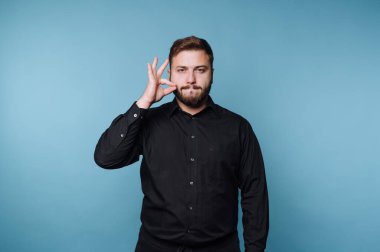 Mavi bir duvarın önünde bir adam duruyor. Yüzünde tarafsız bir ifadeyi tutarken tek eliyle bir el hareketi gösteriyor. Siyah bir gömlek giyiyor..