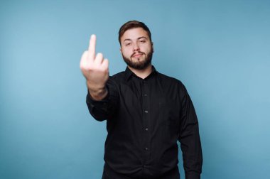 Sakallı bir adam mavi bir arka planın önünde duruyor. O, siyah bir gömlek giyiyor ve orta parmağını kaldırırken ciddi bir ifadeye sahiptir. Şu anda açık bir meydan okuma mesajı var..