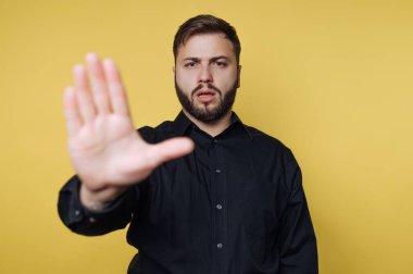 Sakallı bir adam sarı bir duvarın önünde duruyor. Parmakları açık bir şekilde elini kaldırır, ciddi görünür. Bu jest bir duraklama ya da ilgi istiyor gibi görünüyor..