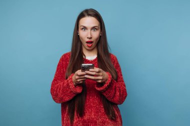 Mavi bir duvarın önünde duran genç bir kadın elinde bir akıllı telefonu tutarken şaşkın şaşkın bakıyor. Onun uzun saçı ve kırmızı kazağı sahnede dikkat çekiyor..