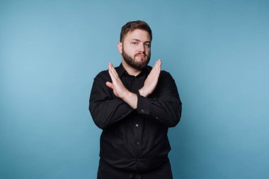 Bir adam mavi arka planda duruyor ve işaret vermek için el hareketi yapıyor. Siyah bir gömlek giyerken odaklanmış ve ciddi görünüyor. Duruşu bir mesaj ilettiğini gösteriyor..