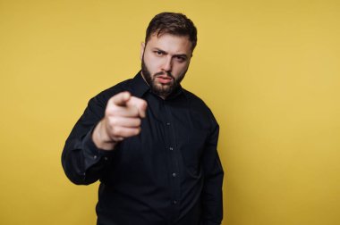 Stüdyoda siyah gömlekli bir adam duruyor. Ciddi bir ifadeyle doğrudan kamerayı işaret ediyor. Arka plan düz sarı..