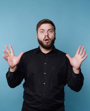 Elleri havada duran bir adam şaşkınlığını gösterir. Şok olmuş gibi bir ifadesi var. Arka plan mavi. Adam siyah bir gömlek giyiyor..