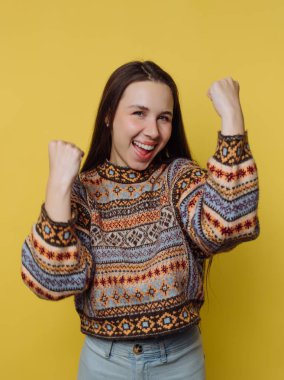 Genç bir kadın yumruklarını kaldırmış ve yüzünde kocaman bir gülümsemeyle ayakta duruyor. Renkli bir kazak ve açık renk pantolon giyiyor. Arka plan parlak sarı, enerjik bir atmosfer yaratıyor..