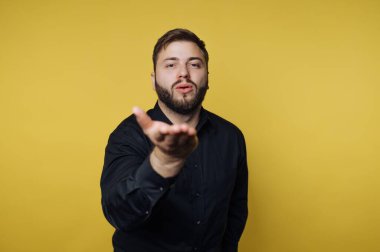 Sarı arka planı olan bir adam öpüşme jesti yaparak elini uzatıyor. Koyu renk bir gömlek giyiyor ve doğrudan seyirciye bakıyor. Ayar parlak ve neşeli görünüyor..