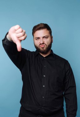 Bir adam ciddi bir ifadeyle mavi bir arkaplanda duruyor. Onaylamadığını işaret ederek baş parmağını aşağı uzatıyor. Siyah gömleği arka planla çelişiyor..