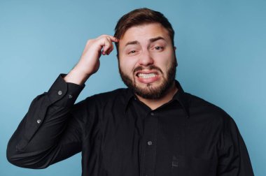 Bir adam kapalı mekanda şaşkın bir ifadeyle kafasını kaşıyor. O, siyah bir gömlek giyiyor ve gün boyunca açık mavi bir duvarın önünde duruyor..