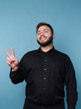 Bir adam gülümseyerek ayakta durur ve sağ eliyle barış işareti yapar. Siyah bir gömlek giyer ve mavi bir duvarın önünde poz verir. Sahne aydınlık ve sıradan..