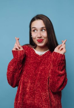 Mavi bir duvarın önünde gülümseyen ve dua eden genç bir kadın. Neşeli bir yüz ifadesi sergiliyor, gündüz aktivitelerinde rahat kırmızı bir kazak giyiyor..