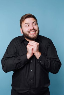 Büyük bir gülümsemeyle, ellerini bir arada tutan bir adam. Heyecanlı ve hevesli görünüyor. Arka plan mavi, neşeli bir atmosfer yaratıyor. Bu sahne neşe ve beklenti gösteriyor..
