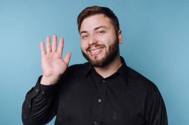 Mavi arka planı olan bir adam içeride duruyor. Kameraya el sallıyor ve gülümsüyor. Siyah bir gömlek giyiyor. Öğleden sonra sıradan bir ayara benziyor..