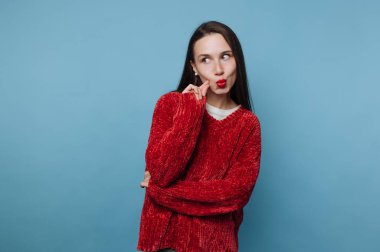 Genç bir kadın kollarını kavuşturup neşeli bir ifade takınıyor. Onun uzun saçı var ve kırmızı bir kazak giyiyor. Arkaplan koyu mavi renkte..