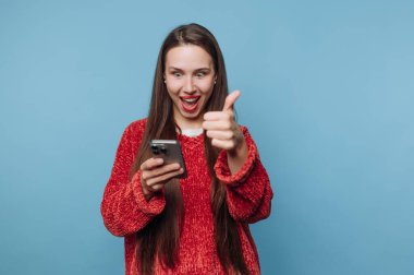 Uzun saçlı genç bir kadın akıllı telefonuna bakar, geniş bir gülümseme içinde. Kırmızı bir kazak giyerken onay işareti olarak baş parmağını kaldırıyor. Arka plan mavi..