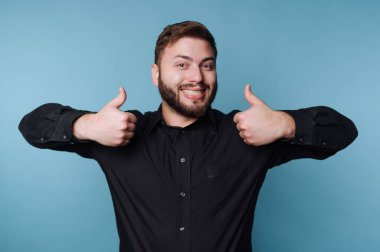 Bir adam mavi bir arka planın önünde duruyor. O, geniş bir şekilde gülümsüyor ve iki başparmak işareti yapıyor. Giysileri gündelik ve mutlu görünüyor. Dekor bir stüdyo gibi görünüyor..