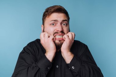 Sakallı bir adam ellerini yüzüne dayayıp endişe ve korku ifade ediyor. Açık mavi bir arkaplanda duruyor, yoğun duygular gösteriyor..