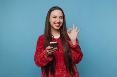 Elinde akıllı bir telefonla duran genç bir kadın. Gülümsüyor ve diğer eliyle iyi bir jest yapıyor. Arka plan basit bir mavi renktir..