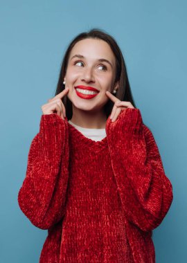 Bir kadın gülümser ve mavi bir arka fonun önünde kırmızı bir kazak giyerken yanaklarını işaret eder. Neşeli ifadesi neşe ve heyecanı akla getiriyor..
