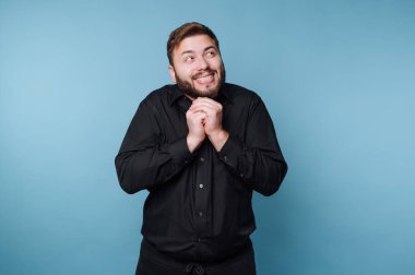 Bir adam açık mavi bir arkaplanda neşeli bir ifadeyle duruyor. Elleri birbirine kenetlenmiş ve yüzünde neşeli bir gülümseme ile coşkulu görünüyor..