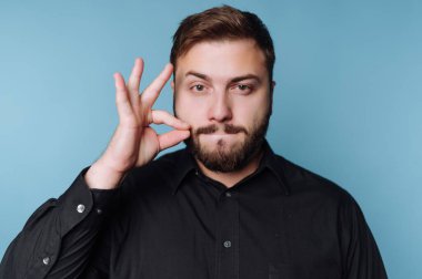 Açık mavi arka planda duran bir adam. Yüzünde ciddi bir ifadeyi korurken bir elini özel bir hareketle havaya kaldırıyor..