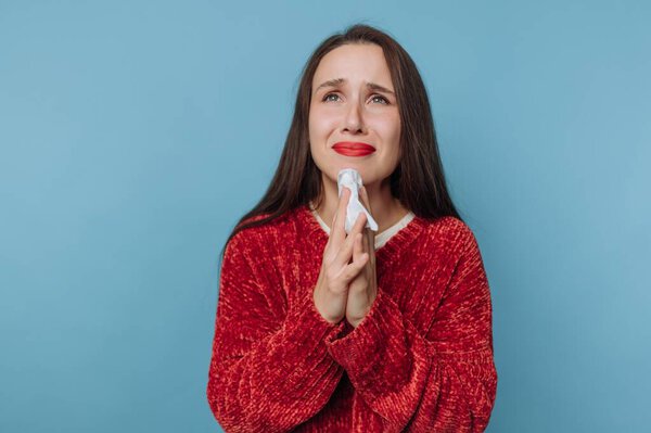 A woman in a red sweater stands against a blue background. She holds a tissue with both hands and shows signs of distress. Her expression indicates she may be feeling emotional or sad.
