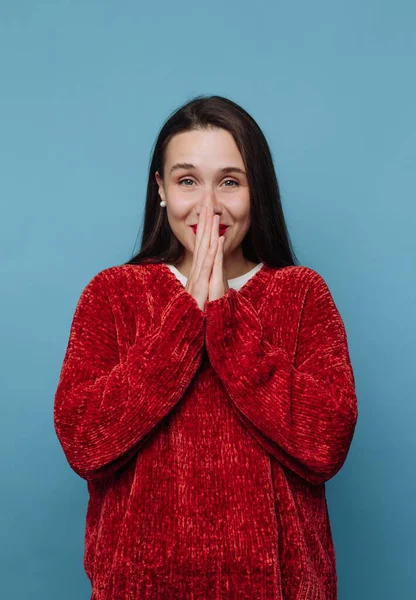 Elleri birbirine bastırılmış ve yüzünde bir gülümsemeyle bir kadın ayakta duruyor. Uzun siyah saçları var ve kırmızı bir kazak giyiyor. Arkaplan koyu mavi renkte..