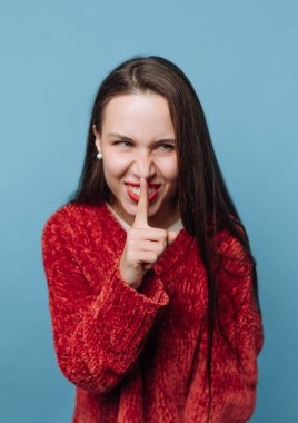 Uzun saçlı bir kadın parmağını dudaklarının üzerine koyarak şşşş hareketi yapıyor. Mavi bir arka plana karşı neşeli bir ifadeyle gülümsüyor..