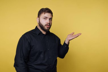 Bir adam parlak sarı bir arka plana karşı duruyor. Siyah bir gömlek giyiyor ve yüzünde şaşırmış bir ifade var. Sorgulama jesti için elini kaldırdı..