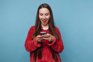 Genç bir kadın mavi bir arka planın önünde duruyor. O bir gülümseme ve parlak kırmızı dudaklarla akıllı telefonuna bakar. Samimi, kırmızı bir kazak giyiyor ve aletle vakit geçirmekten hoşlanıyor..
