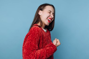 Mavi bir duvarın önünde genç bir kadın duruyor. O, bir yumruk pompasıyla neşeyi ifade eder ve parlak bir şekilde gülümser. Kırmızı süveteri, o anın tadını çıkarırken arka planla çelişiyor..