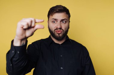 Bir adam sarı bir arka planın önünde duruyor, küçük bedenini göstermek için parmaklarını ayırıyor. Onun şaşırmış bir ifadesi var. Bu sahne kapalı bir yerde geçiyor..