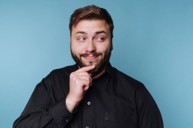 Bir adam düşünceli bir ifadeyle durur, çenesine tek eliyle dokunur. Açık renk sakalı var ve siyah bir gömlek giyiyor. Arka plan mavi..