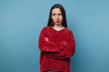 Kolları mavi bir duvara çapraz duran bir kadın. Düşünceli ve biraz da kararsız görünüyor. Onun kırmızı kazağı arka planla çelişiyor. Dekor bir stüdyo..