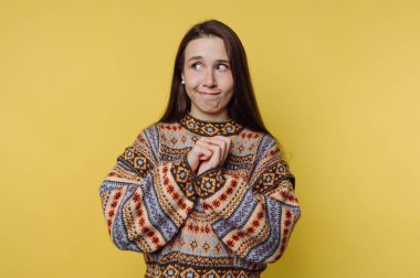 Renkli bir kazak giyen bir kadın ellerini birleştirir. Yüzünde şakacı bir ifade var. Arka plan parlak sarı. Gündüz oldu ve ortam kapalı..