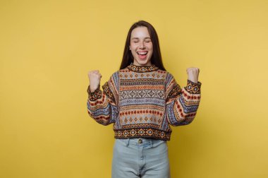 Genç bir kadın büyük bir gülümseme ve sıkılmış yumruklarla ayakta duruyor, heyecan gösteriyor. Parlak sarı bir duvarın önünde renkli bir kazak ve kot pantolon giyiyor..