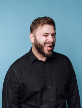 Bir adam mavi bir arka planın önünde duruyor. Siyah bir gömlek giyer ve kameraya göz kırparken gülümser. Yüzünde dostluk ve neşe ifadesi var..