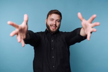 Kolları önde duran bir adam kameraya gülümsüyor. Siyah bir gömlek giyiyor ve mavi arka planı olan bir stüdyoda. Sahne, dostça bir etkileşim olduğunu gösteriyor..