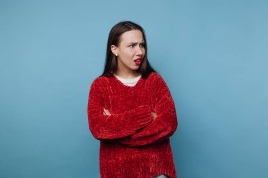 Genç bir kadın kollarını kavuşturup hayal kırıklığını ifade ediyor. Kırmızı bir kazak giyiyor ve uzun koyu renk saçları var. Arkaplan koyu mavi renkte..
