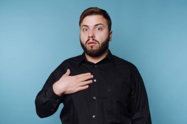 Bir adam bir eli göğsünde ve yüzünde şaşırmış bir ifadeyle ayakta duruyor. Siyah bir gömlek giyiyor ve beklenmedik haberlere tepki veriyor gibi görünüyor. Arkaplan mavi ve sade.