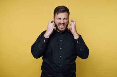 Arkasında sarı bir duvar olan bir adam duruyor. Ellerini başının yanında tutarak hayal kırıklığı belirtileri gösteriyor. Duygularıyla boğuşurken ifadesi güçlü bir duyguyu gösteriyor..
