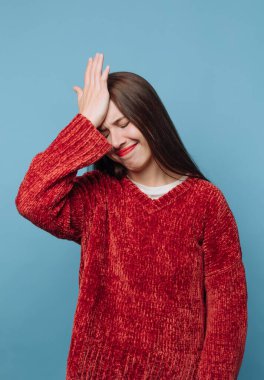 Uzun saçlı bir kadın mavi bir duvarın karşısında duruyor. Kırmızı bir kazak giyiyor ve hayal kırıklığına uğramış görünüyor. Duygularını gösterirken eli alnında duruyor..