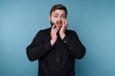 Bir adam endişeli bir yüz ifadesi gösteren mavi bir arkaplan karşısında duruyor. İki eliyle yüzünü tutuyor, o anda bir sıkıntı ya da tedirginlik hissediyor..