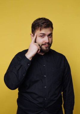 Bir adam parlak sarı bir arka plana karşı duruyor. O siyah bir gömlek giyer ve düşünceli görünürken şakağına dokunur. Dekor kapalı gibi görünüyor..