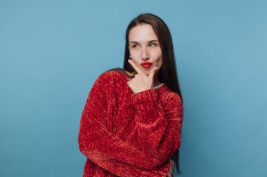 Genç bir kadın elinde kırmızı bir kazakla neşeli bir jest yaparak neşeli bir ifadeyle ayakta duruyor. Mavi arkaplan onun görünümüne karşıtlık ekler.