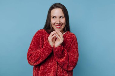 Mavi bir duvarın önünde kırmızı bir kazak giyen bir kadın duruyor. Yüzünde neşeli bir ifade var ve elleri çenesine yakın gülümsüyor..