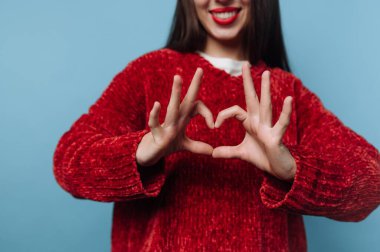 Mavi bir duvarın önünde duran bir kadın elleriyle kalp şekli oluşturuyor. Kırmızı bir kazak giyiyor ve parlak kırmızı dudakları var. Ellerini gösterirken gülümsüyor..
