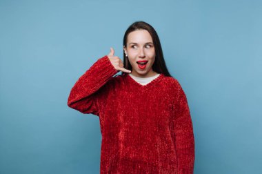 Bir kadın telefon hareketleriyle gülümsüyor. Kırmızı bir kazak giyiyor ve uzun koyu renk saçları var. Arka plan mavidir, sahneyi aydınlık hissettirir..