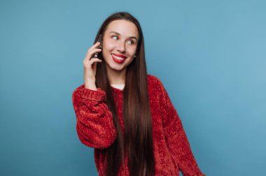 Genç bir kadın telefonuyla konuşurken mavi bir duvarın önünde duruyor. Gülümsüyor ve sohbete dalmış gibi görünüyor. Uzun saçları omuzlarından aşağı akıyor..